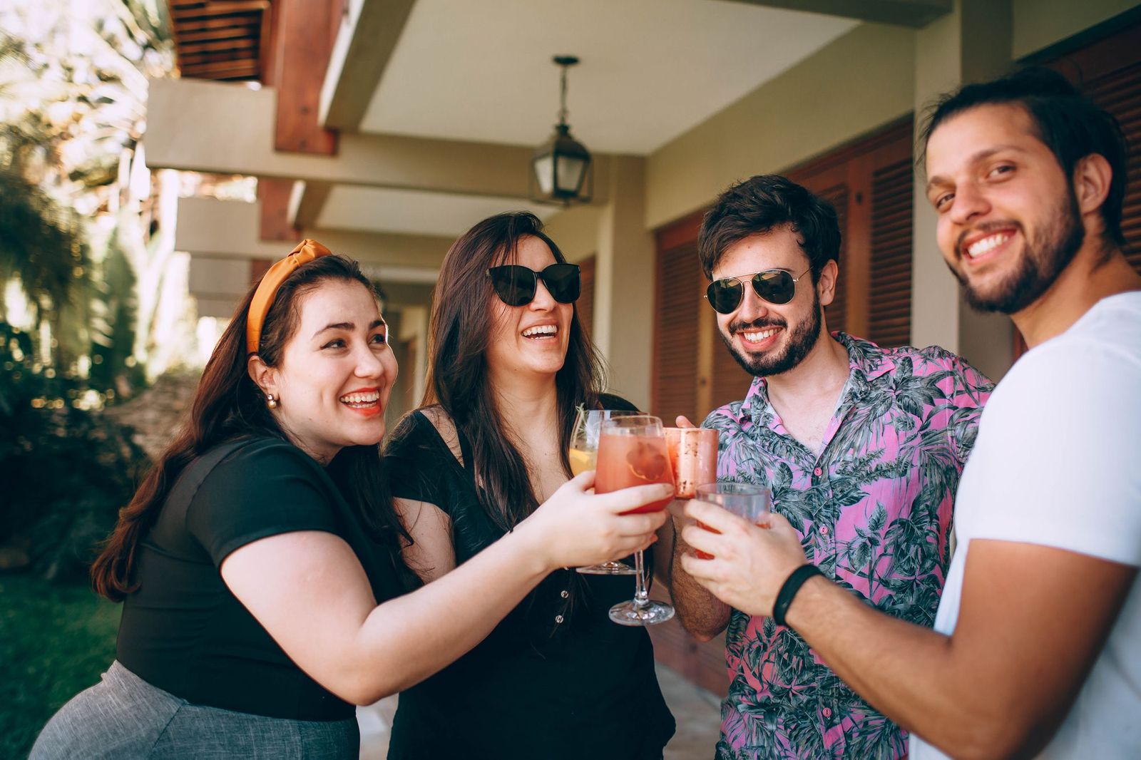 Four friends standing on a shaded porch in the sunshine, laughing and clinking glasses of cocktails together