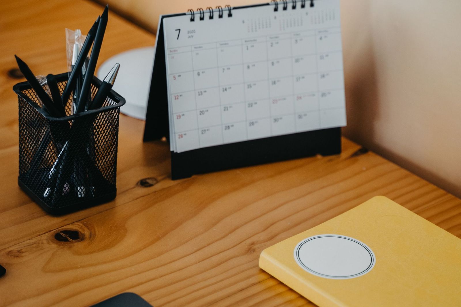 A small black and white desk calendar standing open on a wooden desk beside a mesh holder full of pens and a yellow notebook