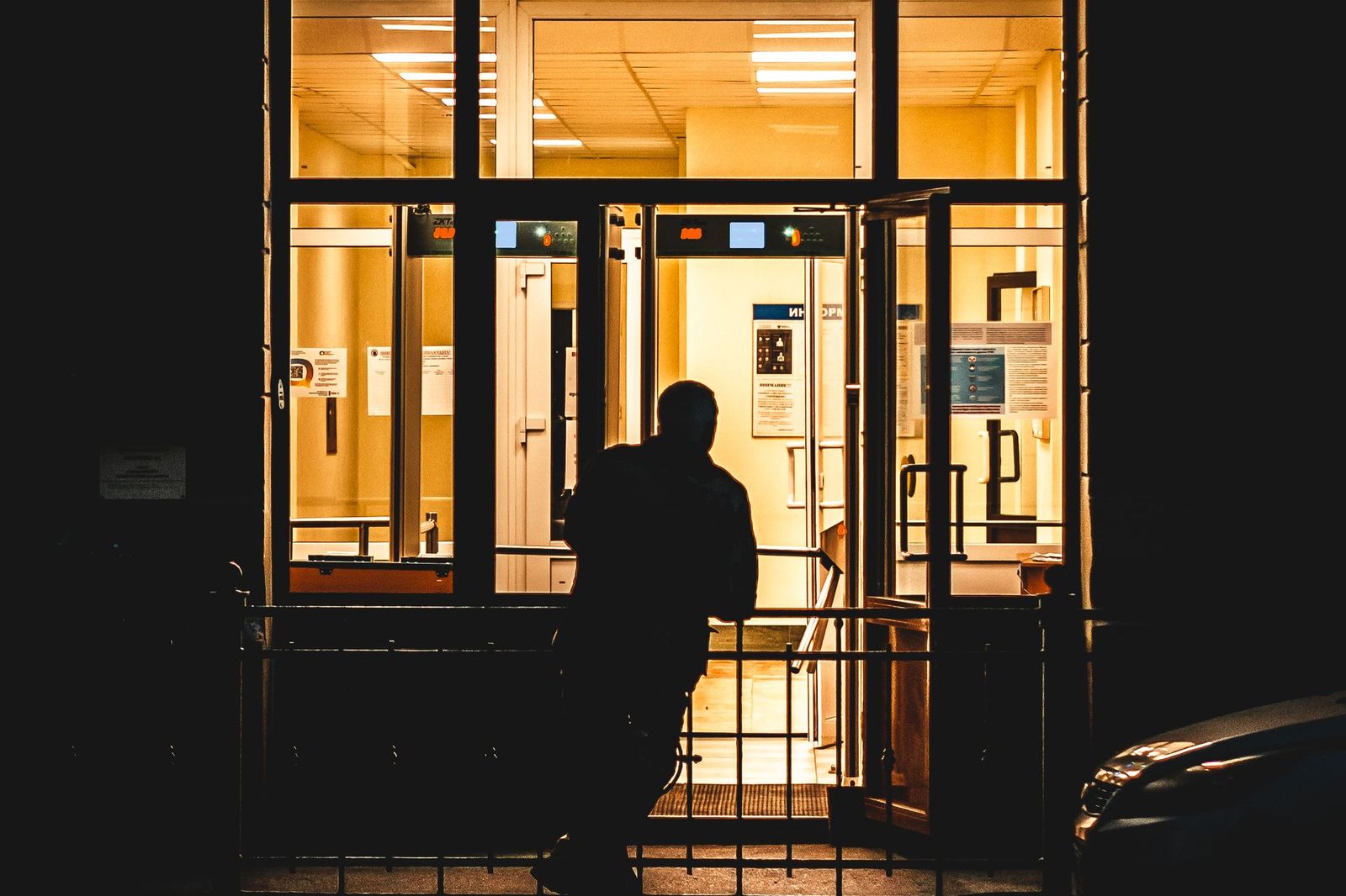 Silhouette of a person walking toward a brightly lit building entrance at night, passing through an open security gate