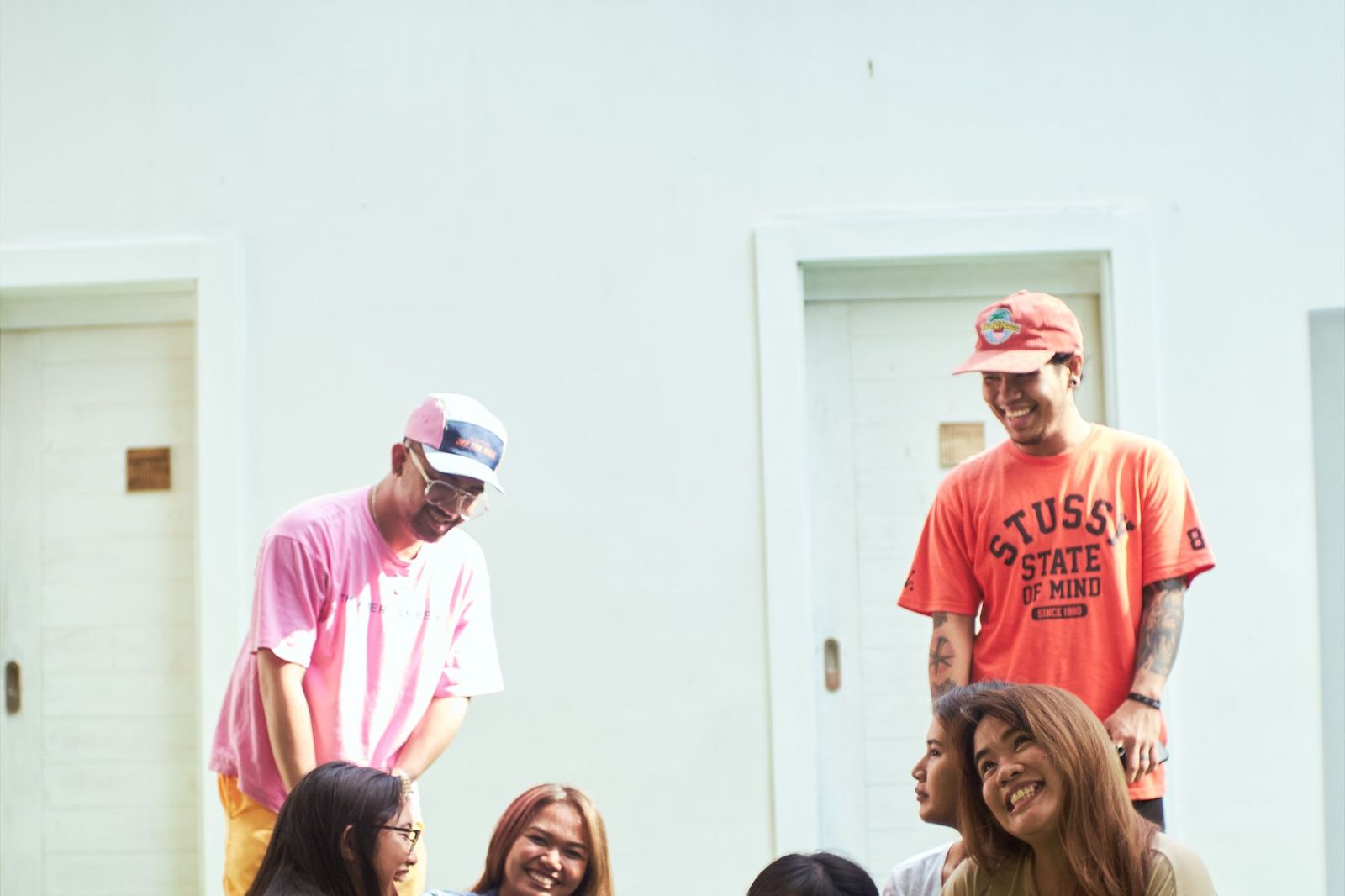 Six friends in bright t-shirts and caps gathering and laughing outside a pale doorway in daylight, some standing and some seated below