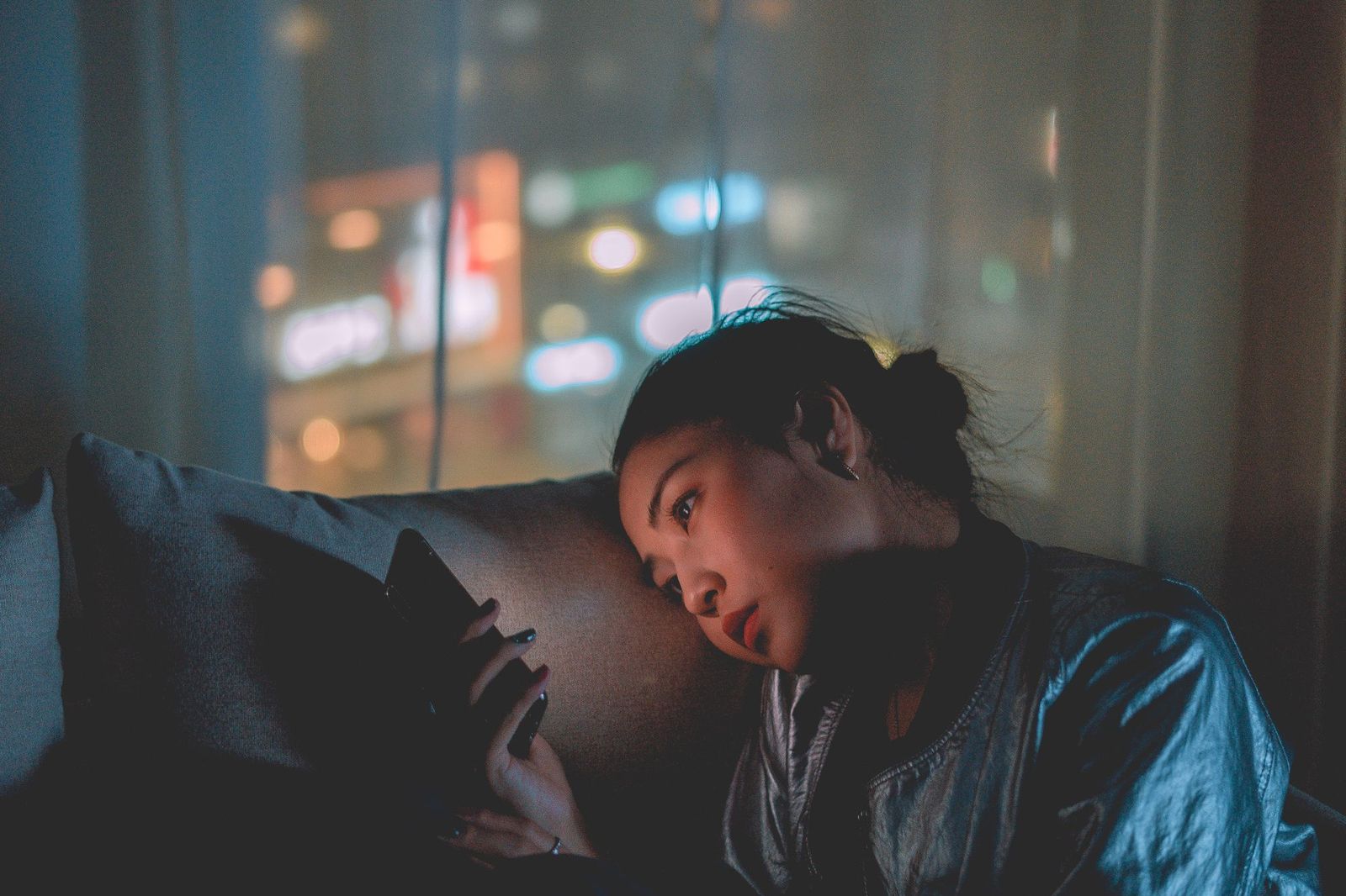 A woman lying against a cushion in a dark room at night, her face lit by her phone screen, city lights blurred in the window behind her