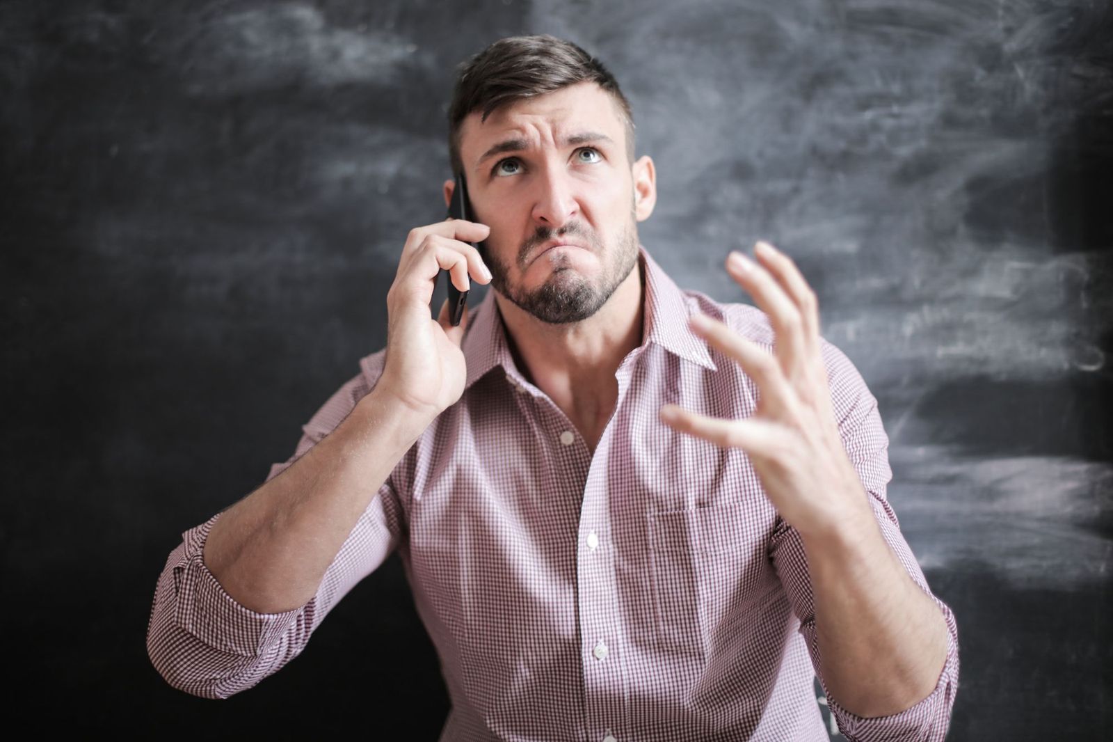 A man in a checked shirt holding a phone to his ear, frowning and gesturing upward with his free hand against a dark chalkboard wall