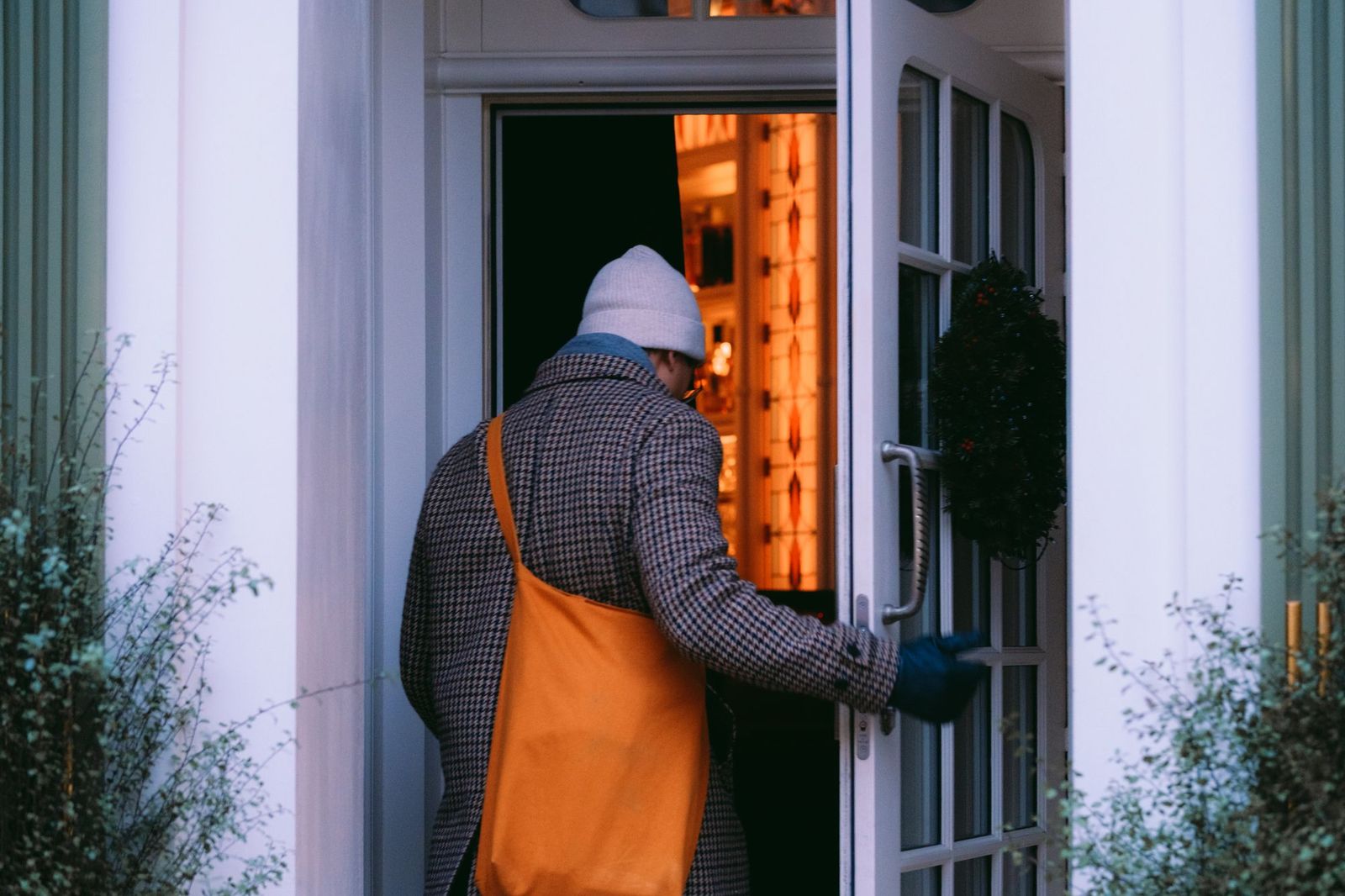 A person in a checked coat and knitted hat pushing open a lit front door at dusk, carrying an orange tote bag over one shoulder