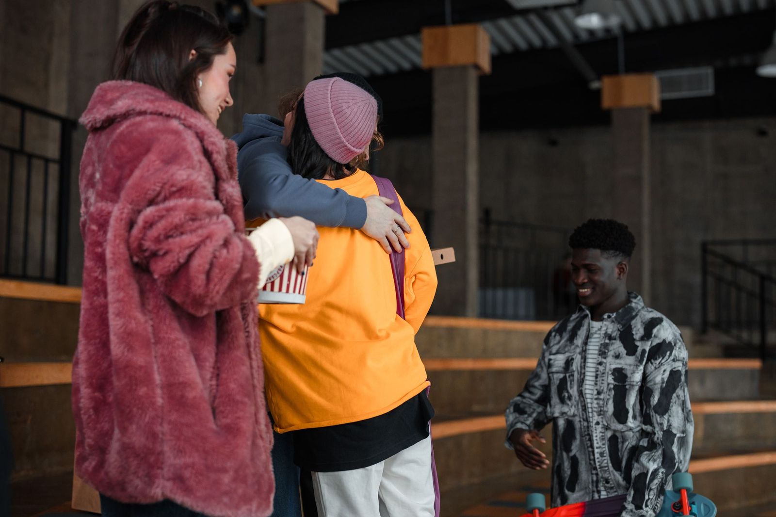 Three friends greeting each other outside a concrete building, two of them hugging while a third arrives smiling with a skateboard
