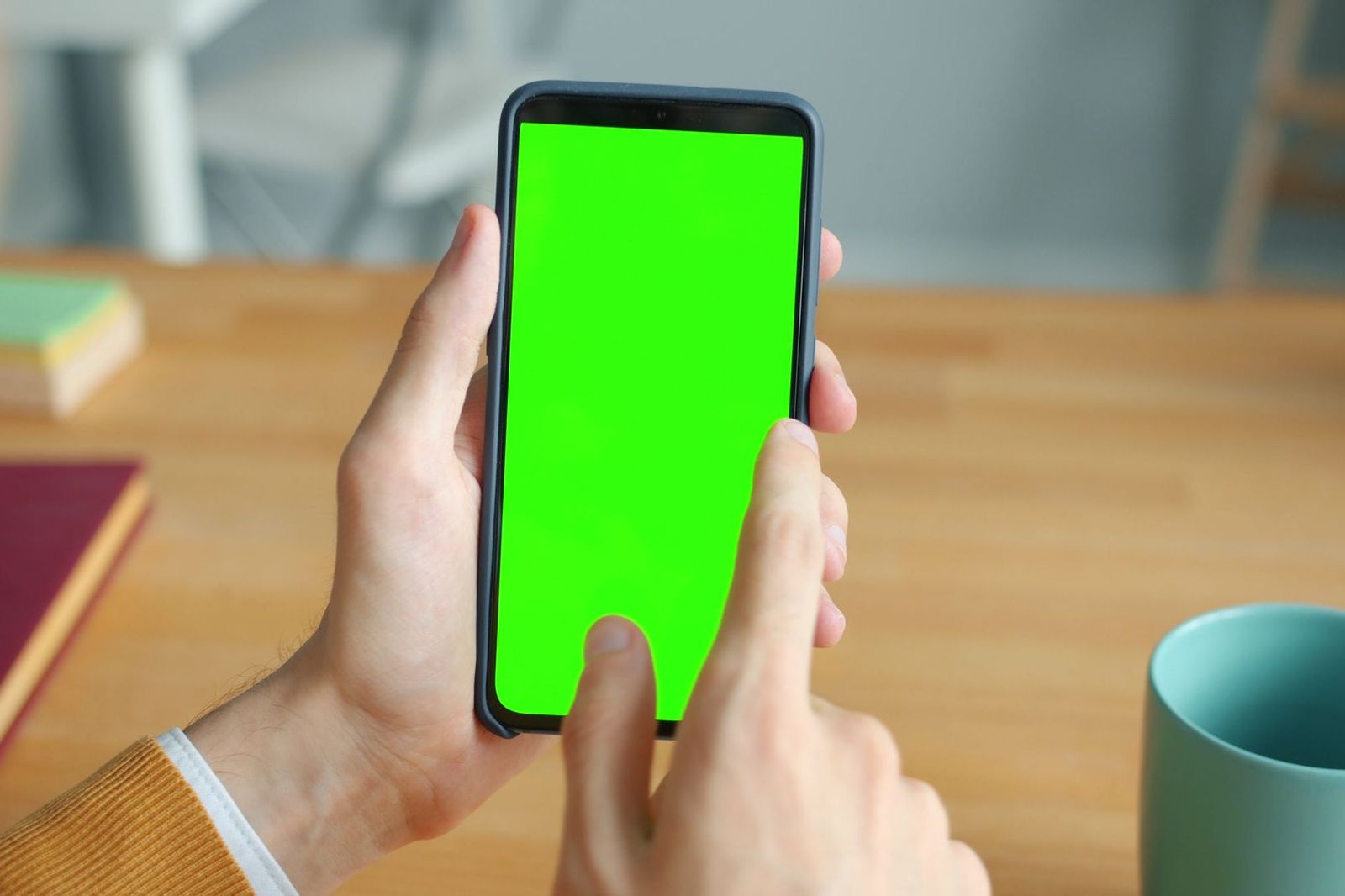 A smartphone with a blank green screen held in two hands over a wooden desk with books and a mug nearby