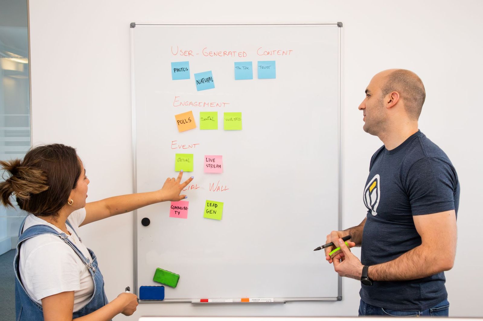 Two people standing at a whiteboard covered in coloured sticky notes, one pointing at a note while the other listens holding a marker
