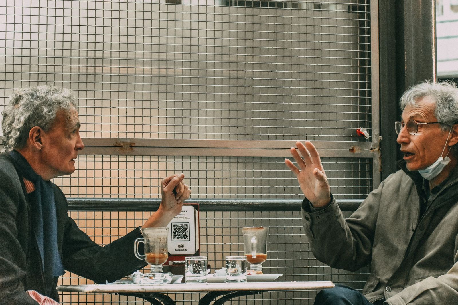 Two older men sitting across a small cafe table outdoors, one gesturing mid-sentence while the other leans in to listen