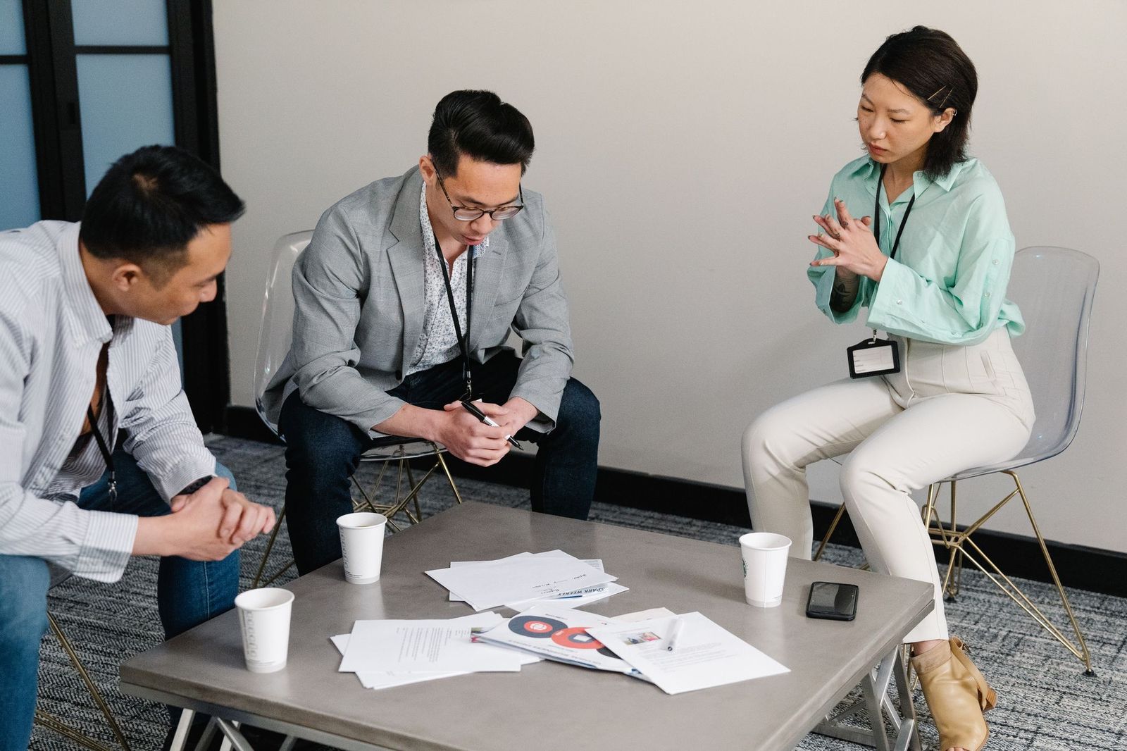 Three colleagues sitting around a low table in a meeting room, papers and coffee cups spread out, one of them talking with her hands
