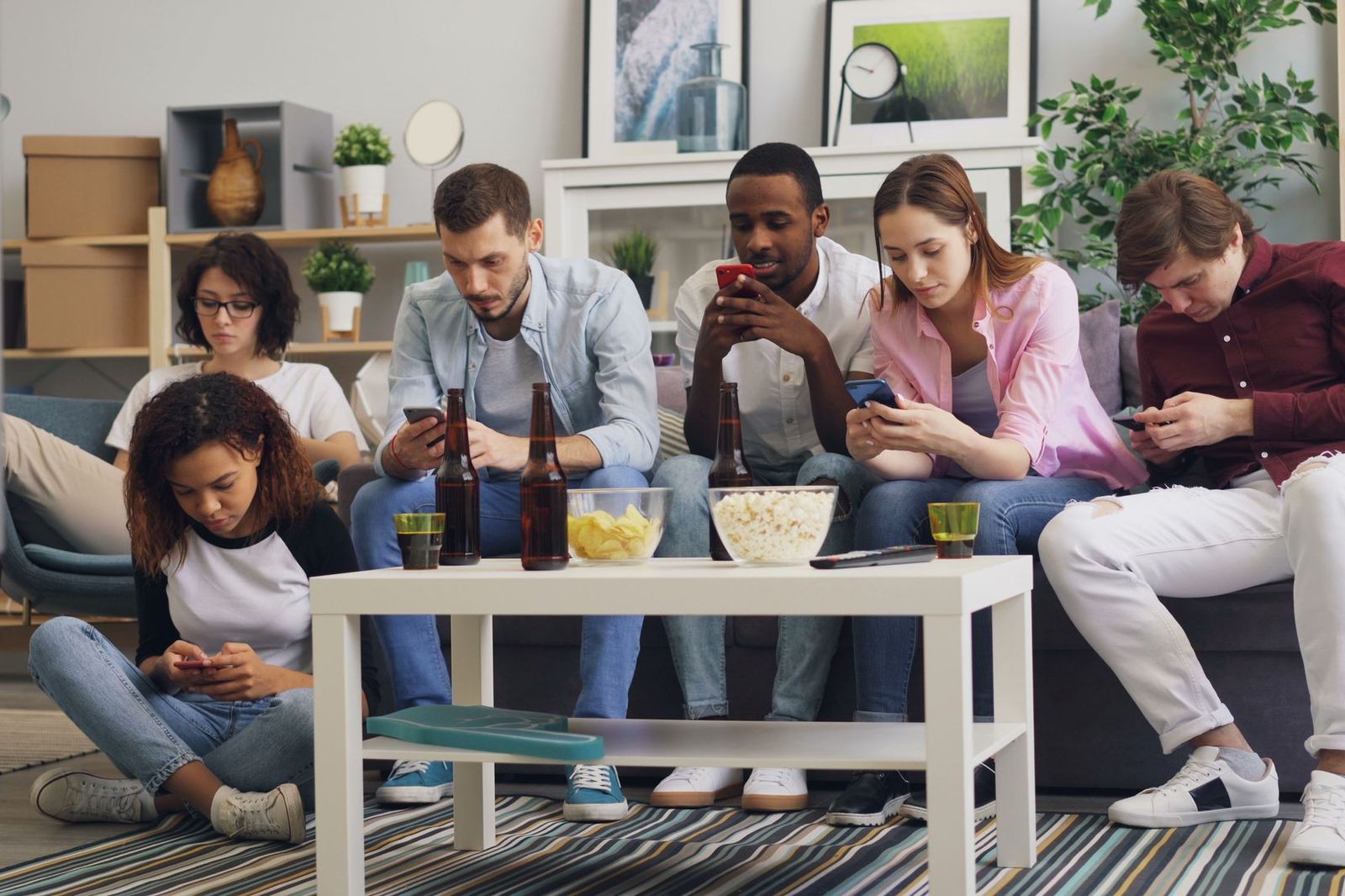 A group of six friends sitting together on a sofa, each looking down at their own phone, with drinks and snacks on the table
