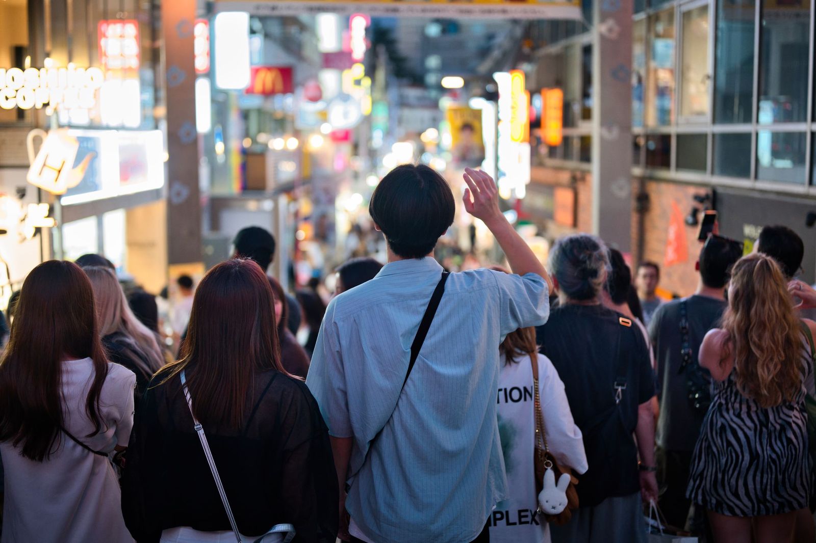 A crowded pedestrian street at dusk with neon signs, people walking together and one person raising a hand in greeting