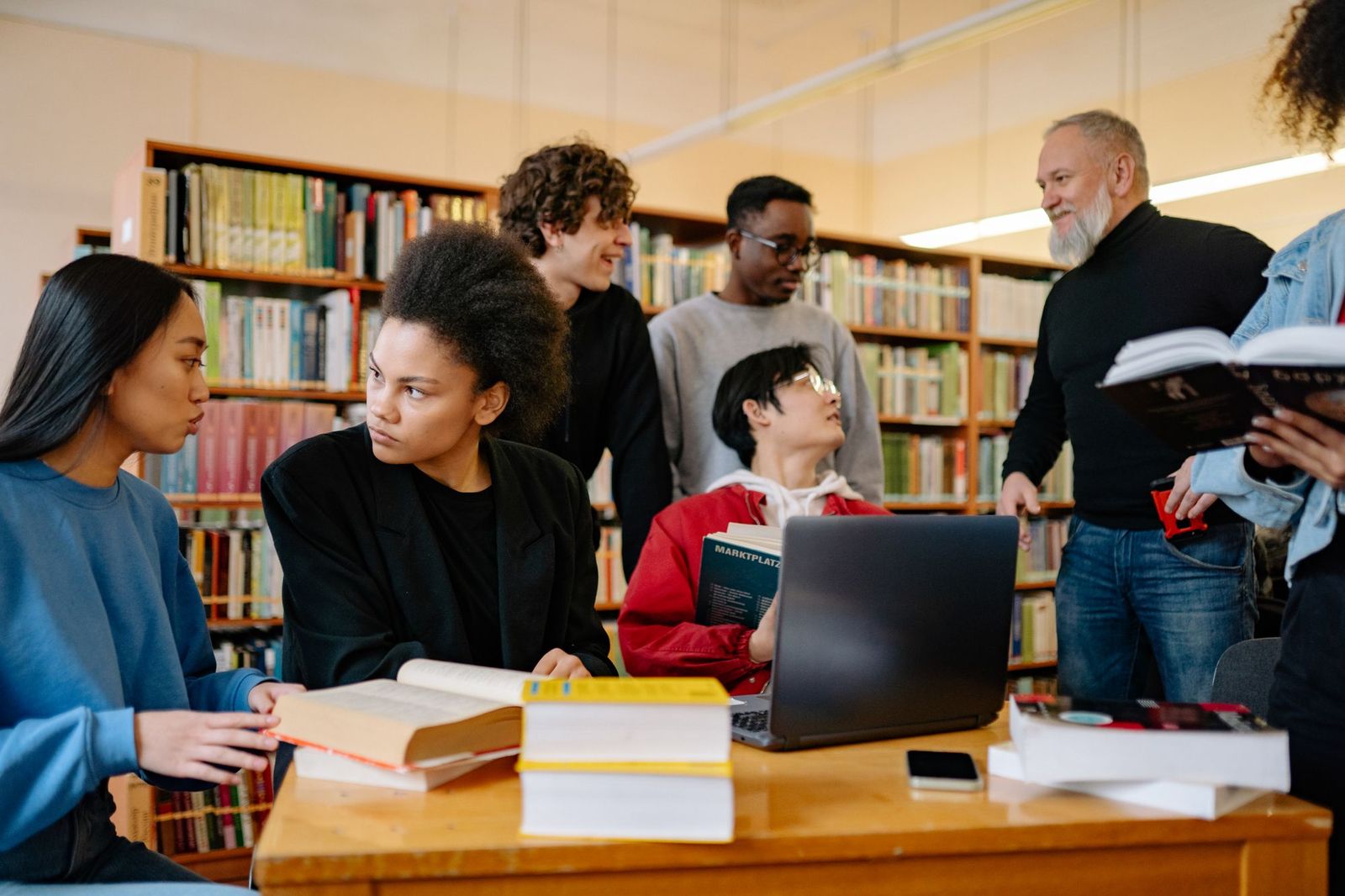 Six students gathered around a library table stacked with books and a laptop, several of them talking at once while an older man leans in from the side