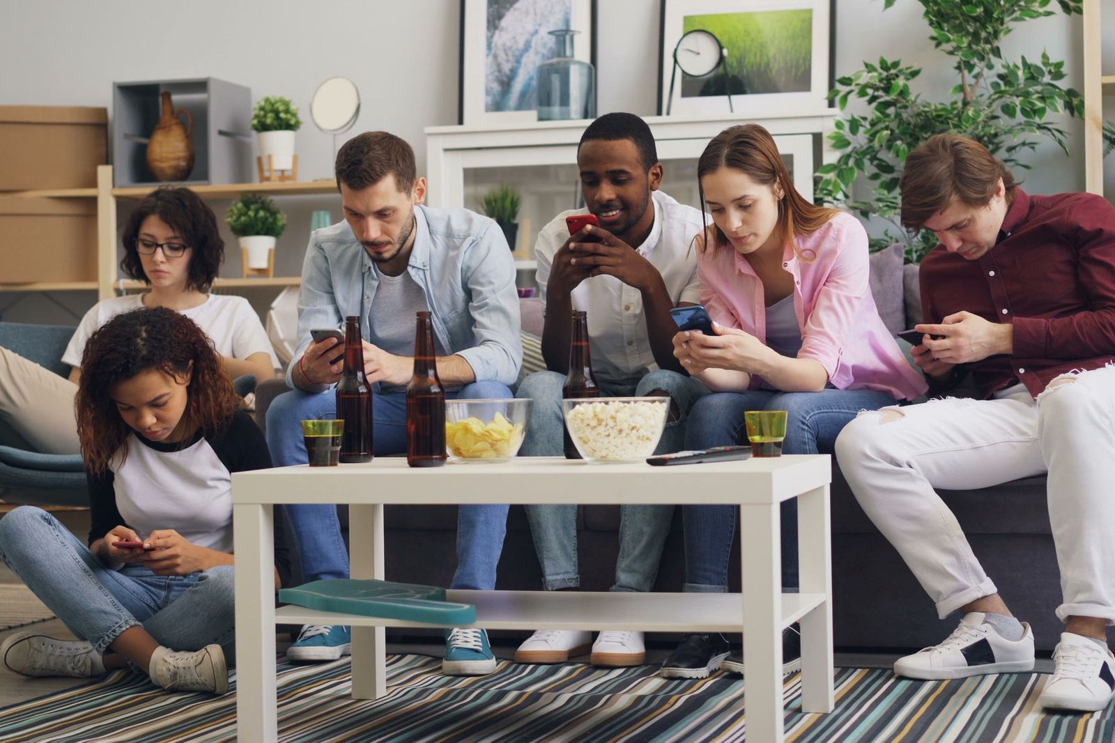 Six friends sitting together on and around a sofa in a living room with snacks on the table, every one of them looking down at their own phone