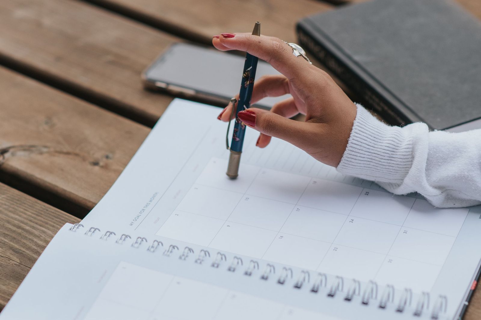 A hand holding a pen over an open spiral-bound monthly planner on a wooden table, with a phone and a closed notebook beside it