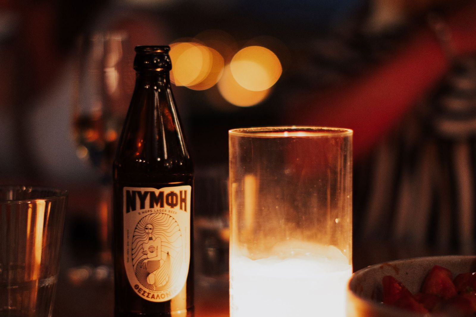 A beer bottle and a lit candle in a glass on a bar table at night, with warm out-of-focus lights behind