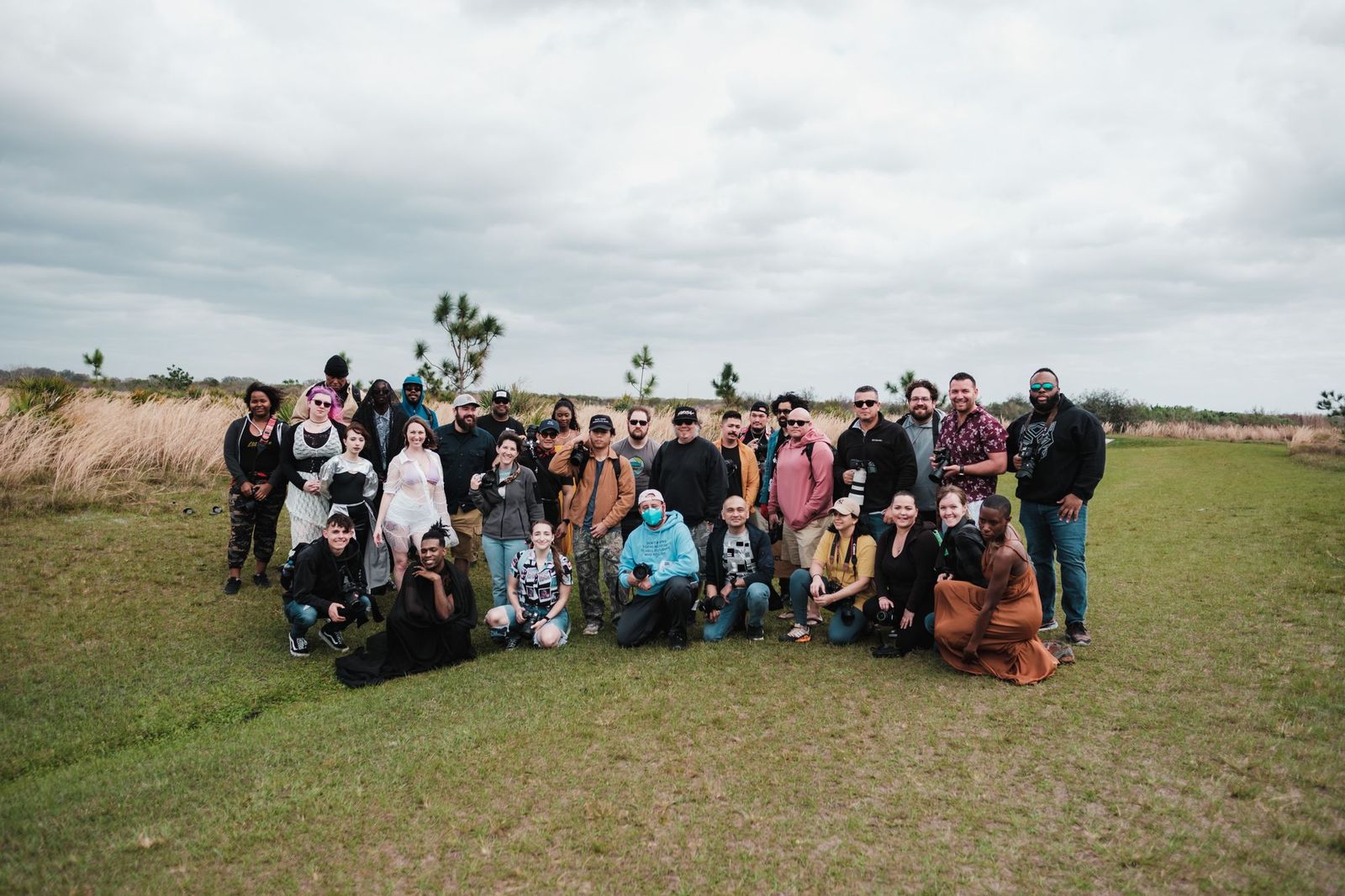 A large group of about thirty people with cameras posing together on grass under a grey sky, some standing and some kneeling in front
