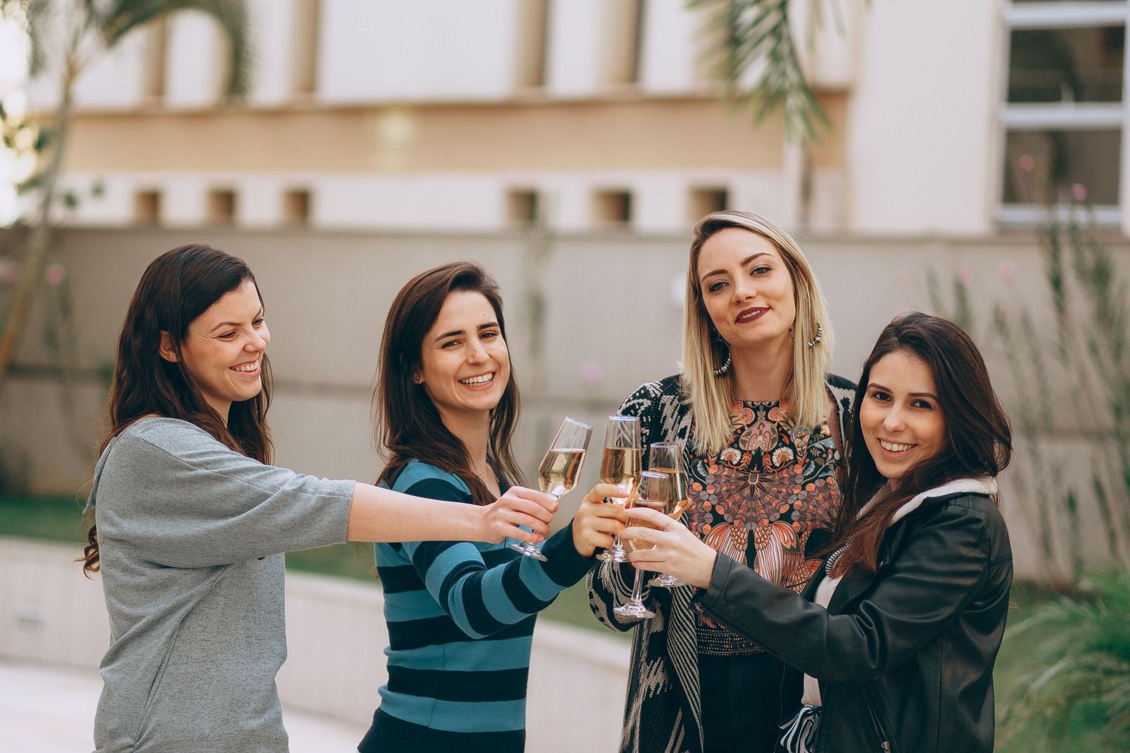Four women standing outdoors in front of a pale building, smiling and touching their glasses of sparkling wine together in a toast