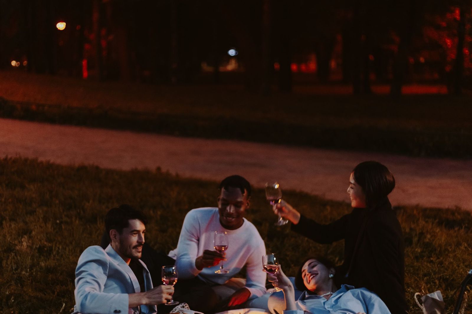Four people sitting on the grass in a park after dark, lit by warm light, holding glasses of wine and laughing together, with a path and trees behind them