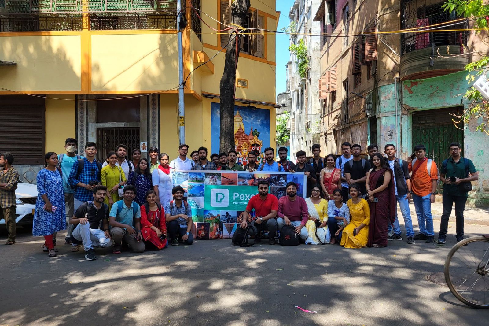 A large group of about forty people posing together on a city street, some standing and some crouching in front, holding a banner between them, with balconies and shuttered shopfronts behind