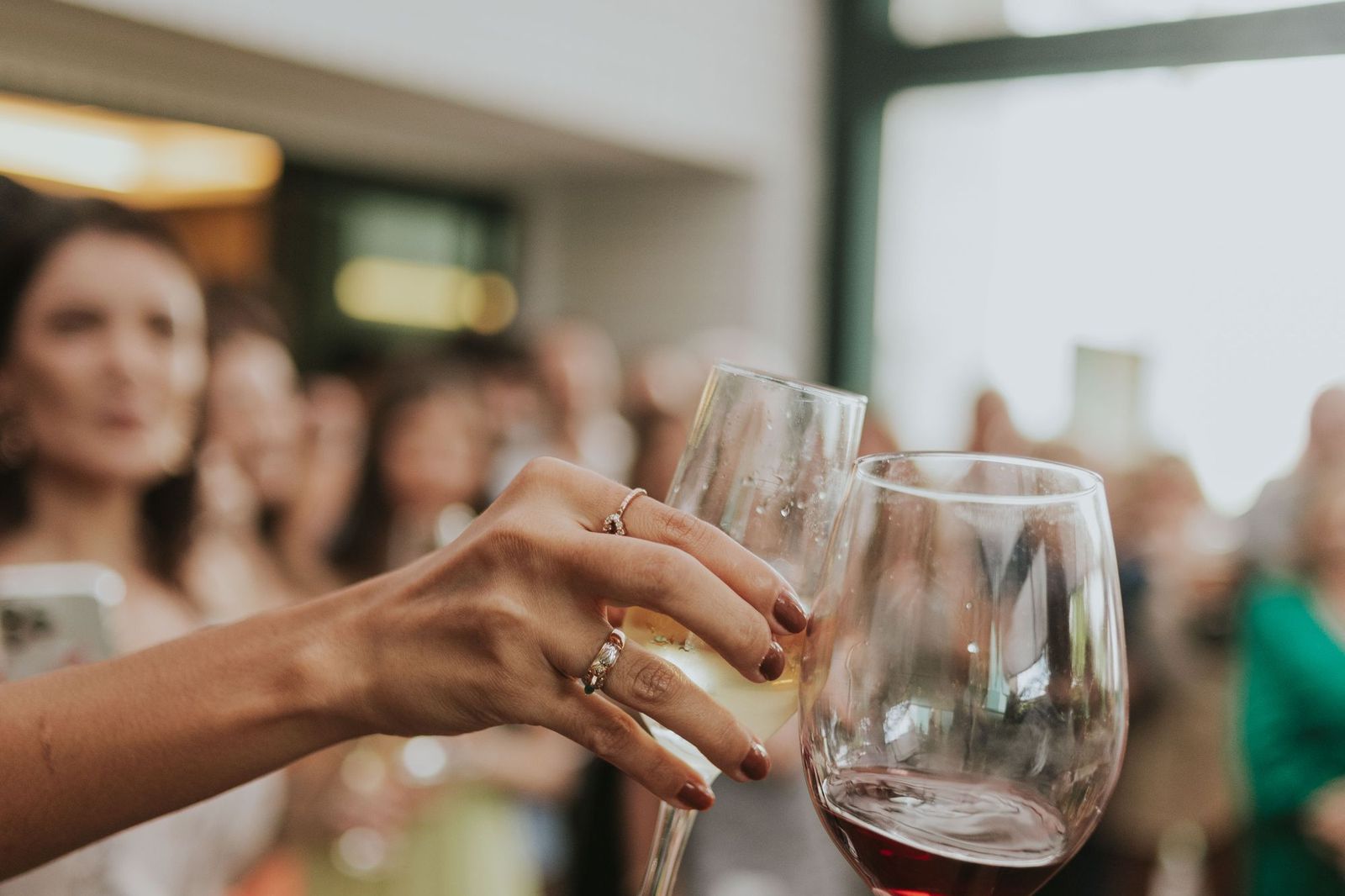 Two hands reaching in to clink a wine glass and a champagne flute together, with a blurred crowd of people behind