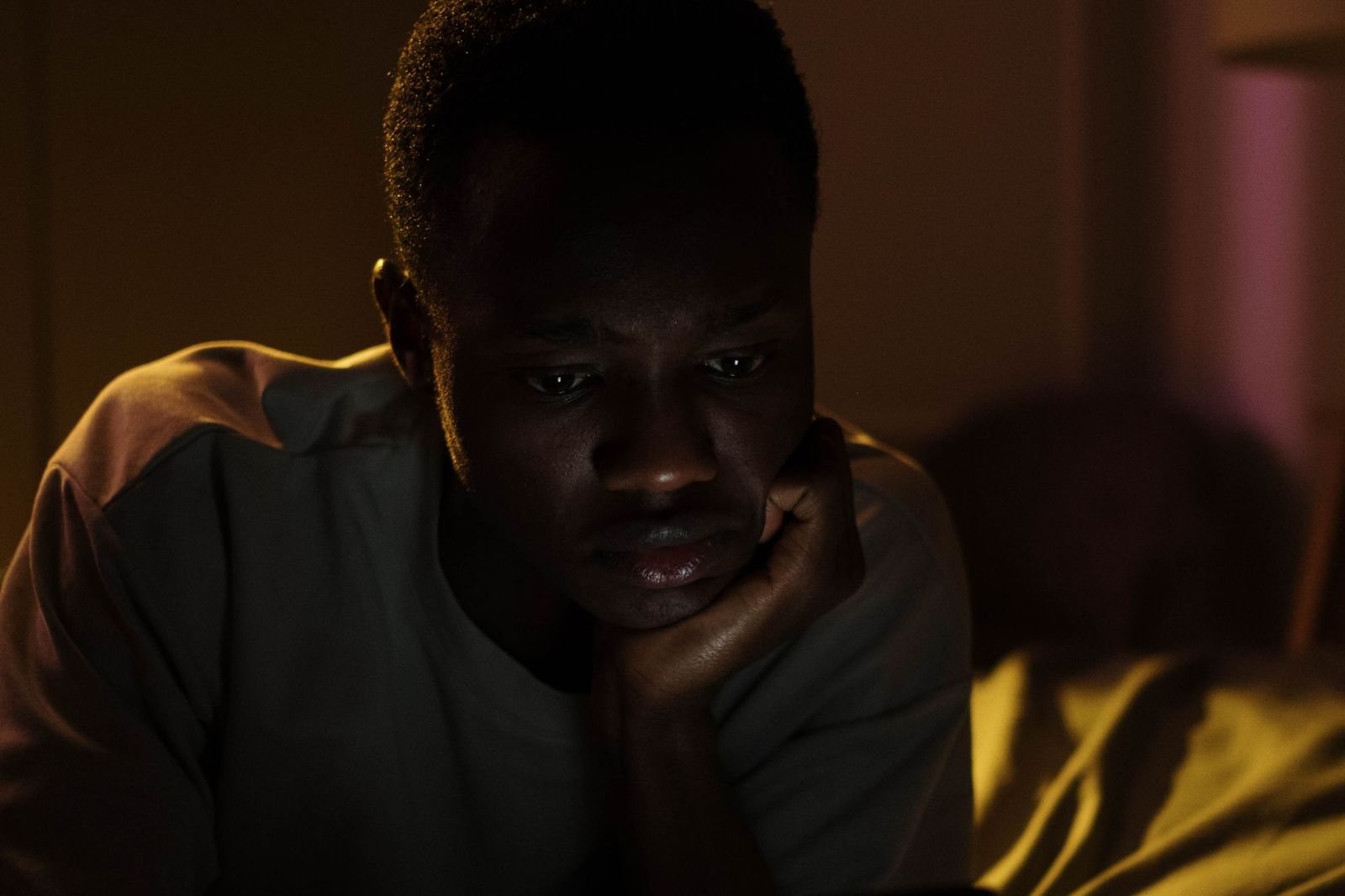 A young man alone in a dark room, chin resting on his hand, face lit from below by a screen as he reads something