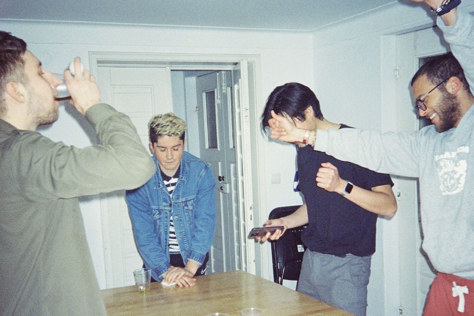 Four friends standing around a kitchen table in a bright room, one drinking, one laughing and shoving another, one holding a phone
