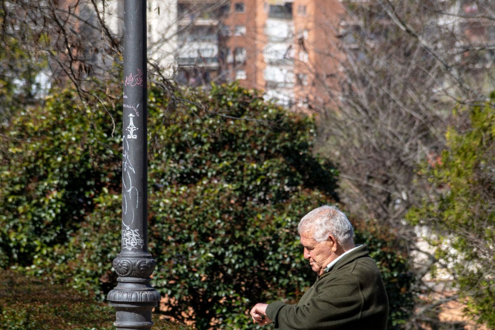 An older man in a green jumper sitting alone beside a lamppost in a city park, looking down at his wrist