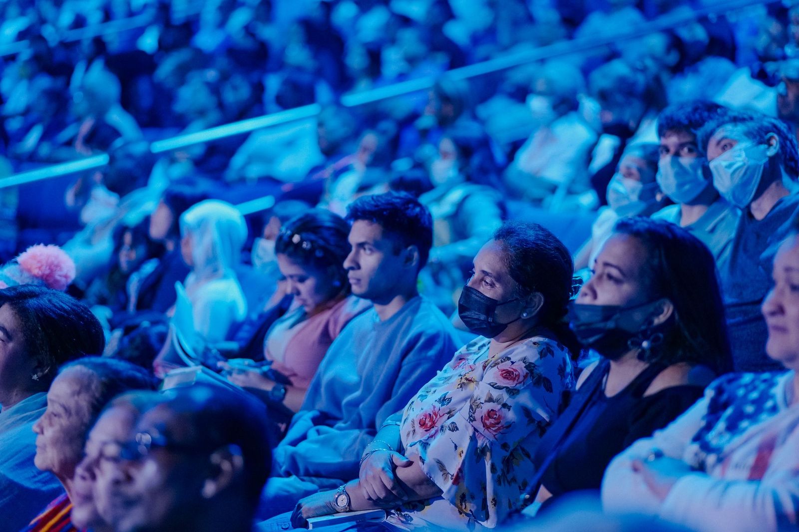 A packed tiered audience at a venue lit in deep blue, rows of seated people watching something out of frame