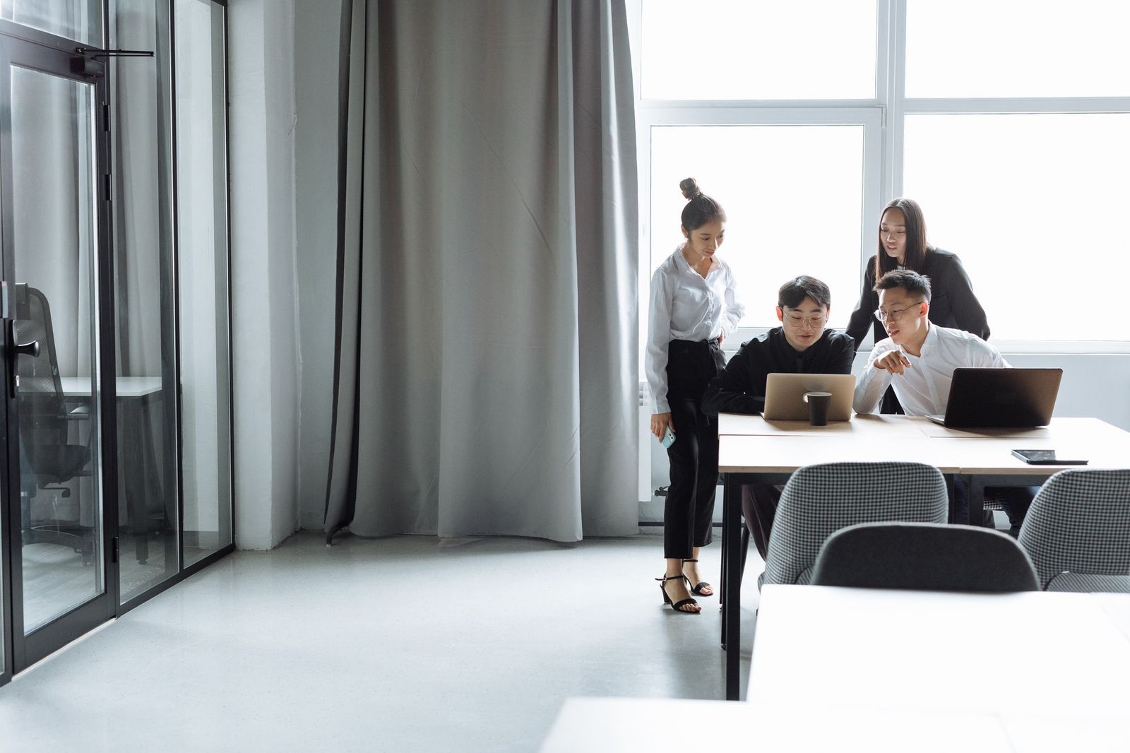 Four colleagues in an open-plan office gathered around two laptops at a long table beside a bright window
