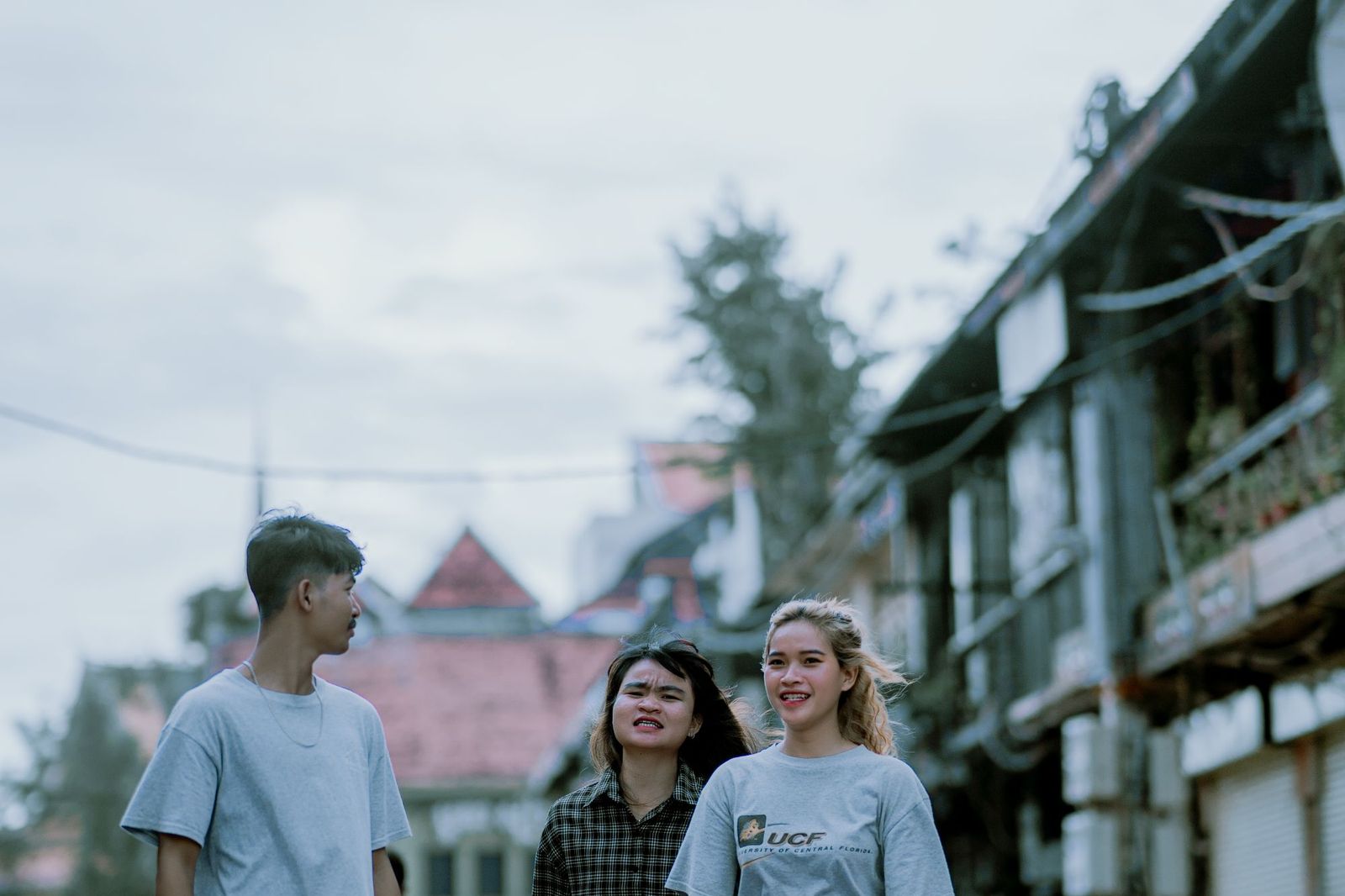 Three young people standing and talking on a street of old wooden shophouses under an overcast sky