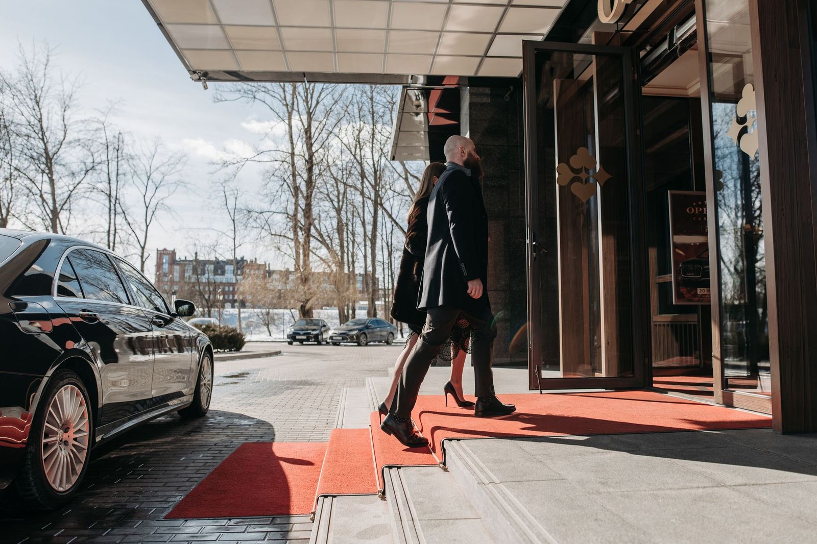 A man in a dark overcoat and a woman walking up red carpeted steps into a glass building entrance on a bright winter day