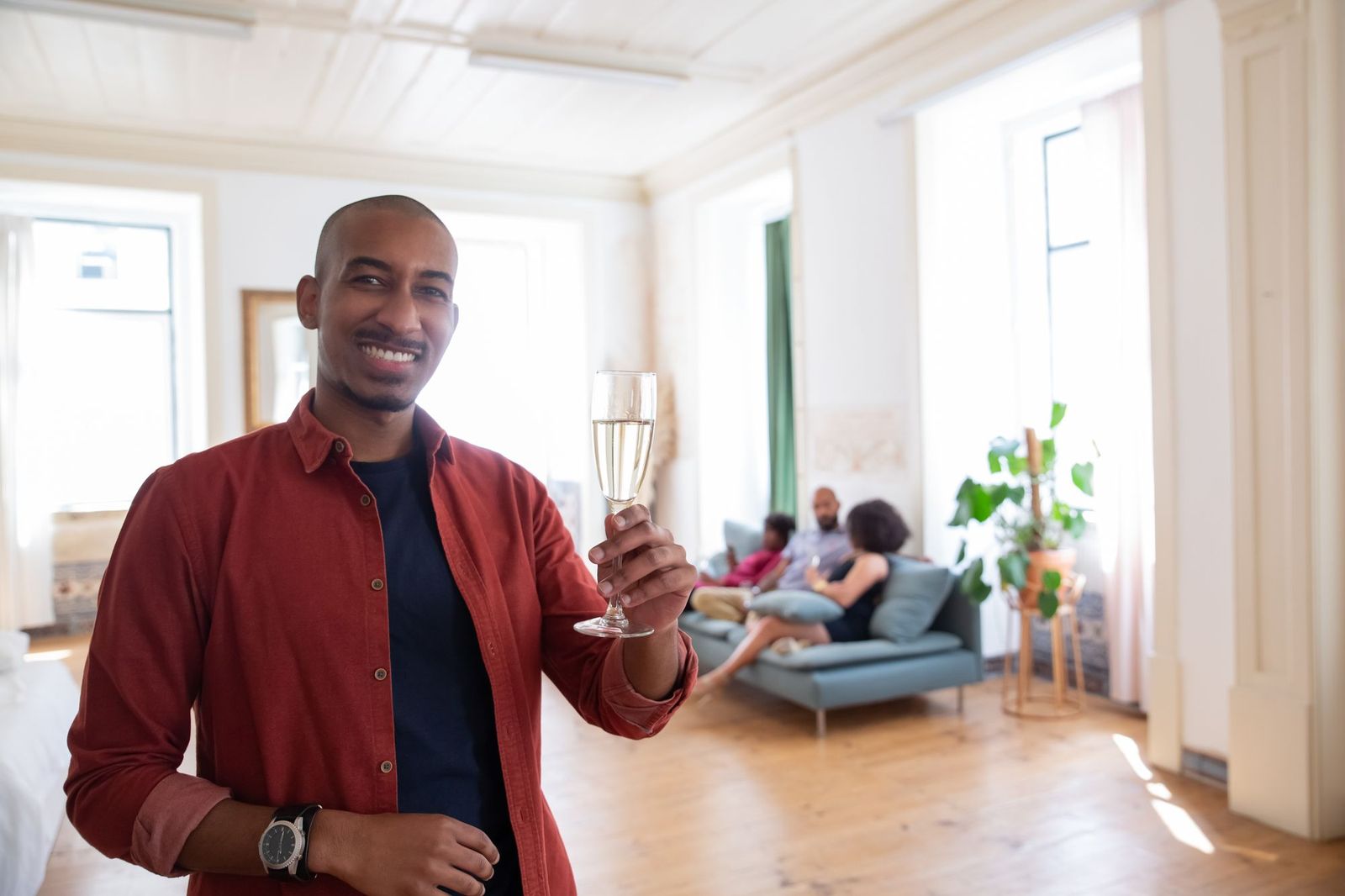 A smiling man in a red shirt holding a glass of sparkling wine in a bright apartment, with three guests sitting on a sofa behind him