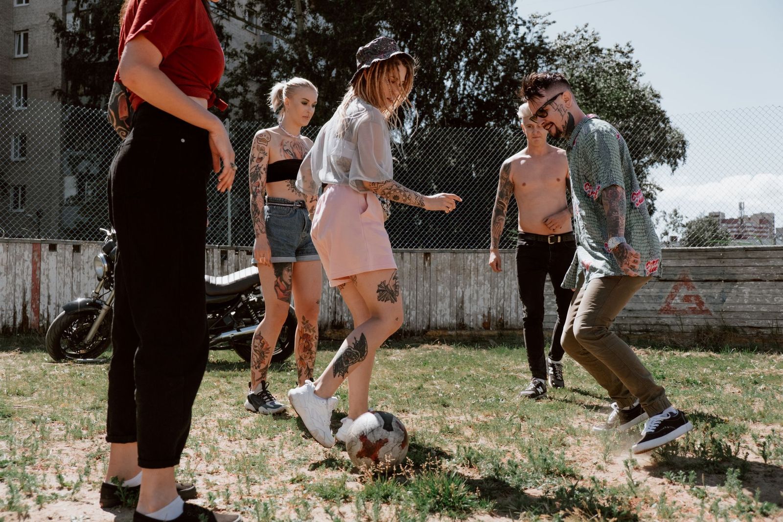 Five friends in casual summer clothes playing football on a patchy grass yard by a chain link fence, one about to touch the ball