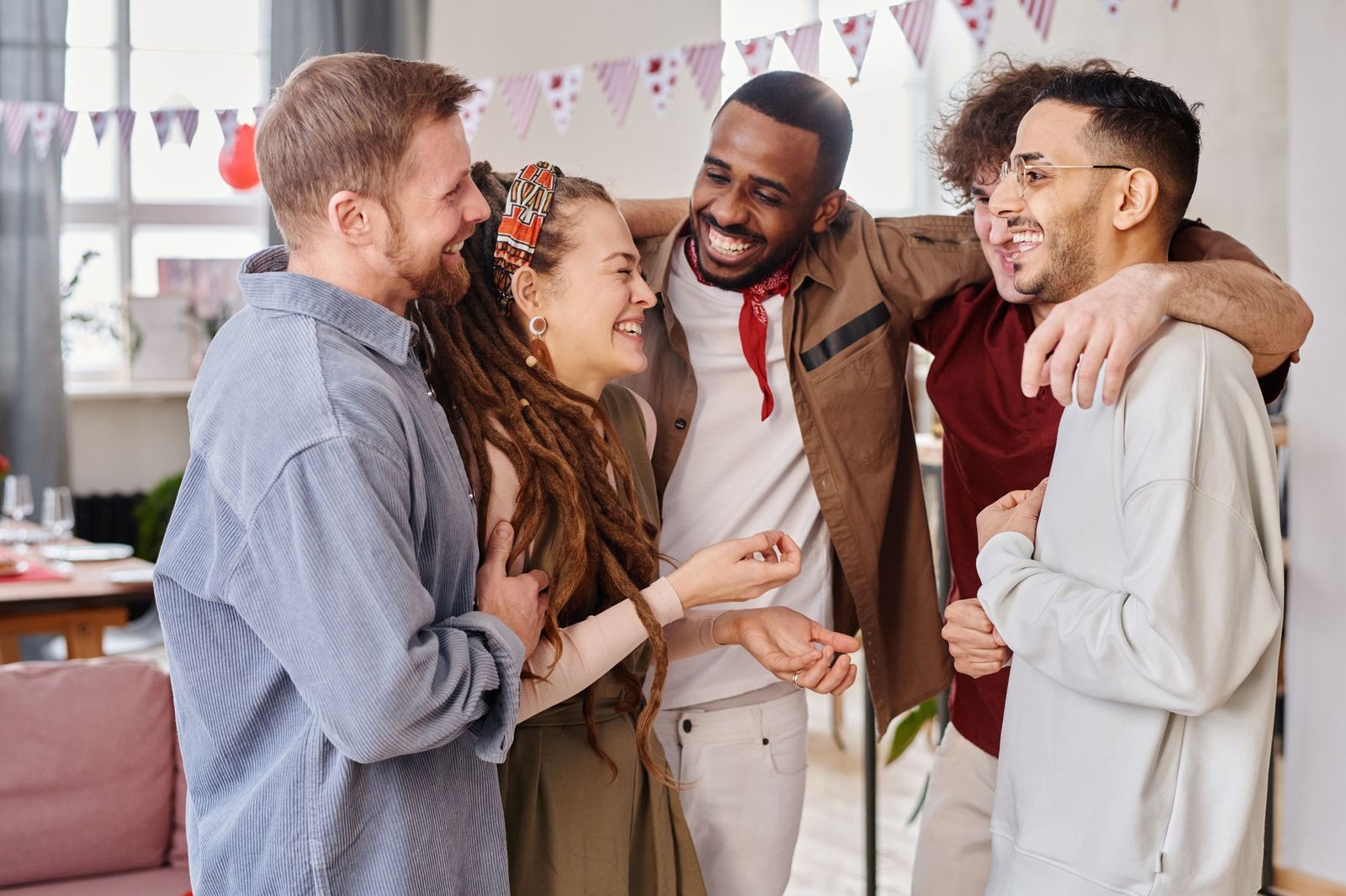 Five friends standing together laughing in a living room decorated with bunting and a balloon, arms around each other's shoulders