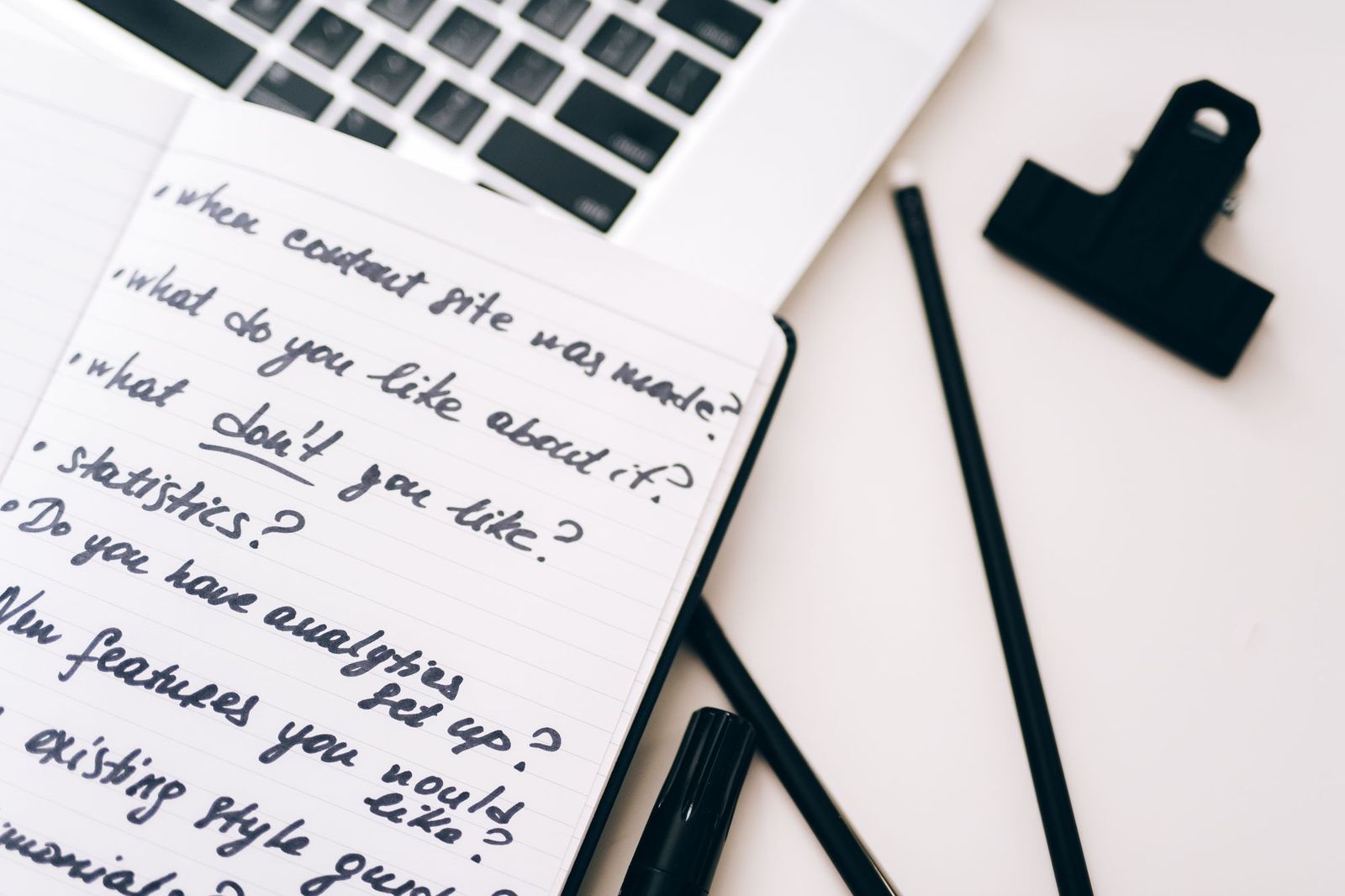 A notepad covered in handwritten questions lying open on a desk beside a laptop keyboard, with two pencils, a marker pen and a black bulldog clip