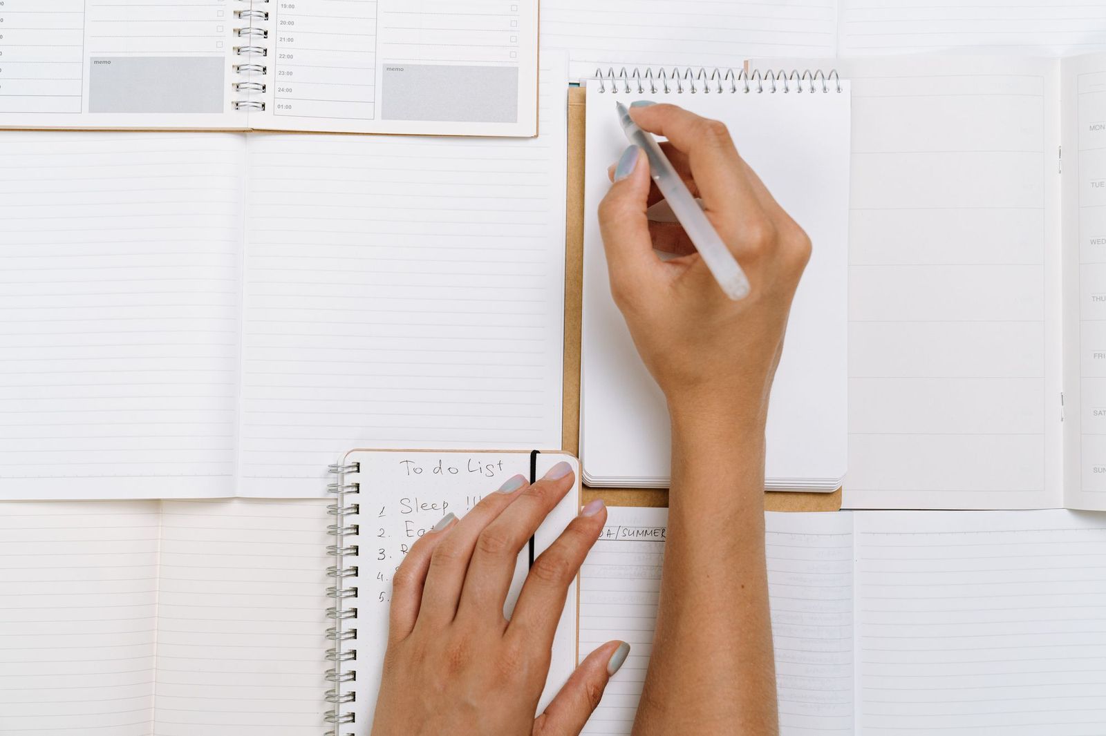 Overhead view of two hands writing on a small spiral notepad, surrounded by open planners and a handwritten to-do list