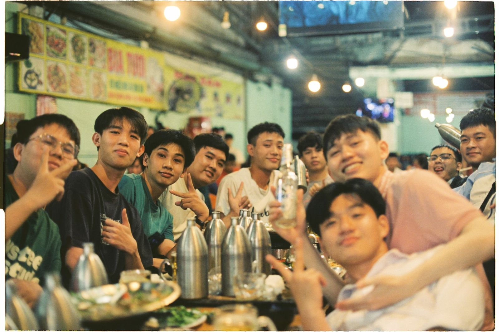 A large group of young friends packed shoulder to shoulder along a table at a busy open-air restaurant at night, smiling and making peace signs over bottles and shared dishes under strings of bulbs