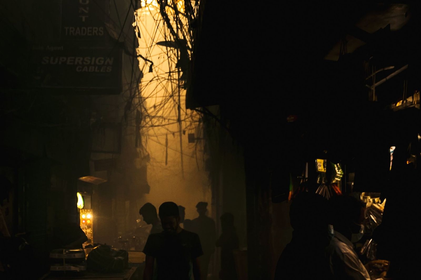 A narrow street at night lit by a single glowing sign, tangled overhead wires, and a few silhouetted figures walking through the smoky light
