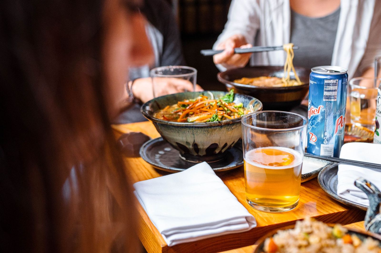Two friends eating noodles and hotpot together at a restaurant table, chopsticks in hand, drinks and side dishes spread across the wood table