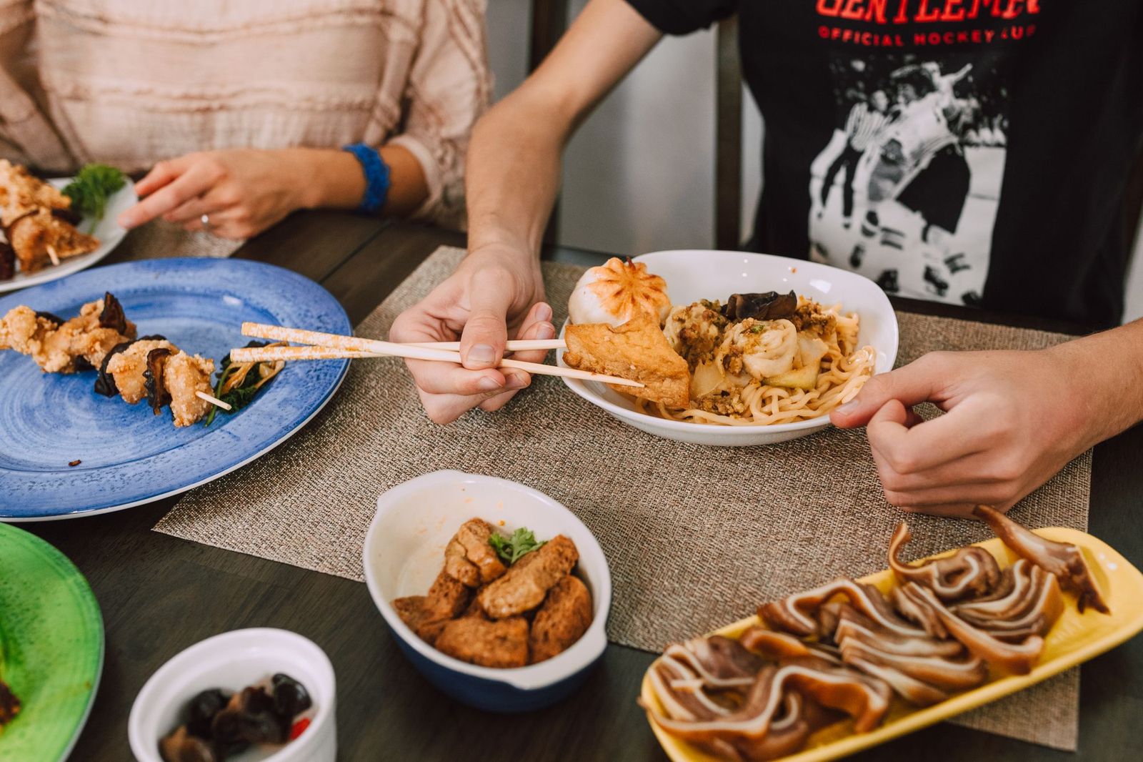 Two people's hands sharing chopsticks over a shared plate of noodles and dumplings at a table