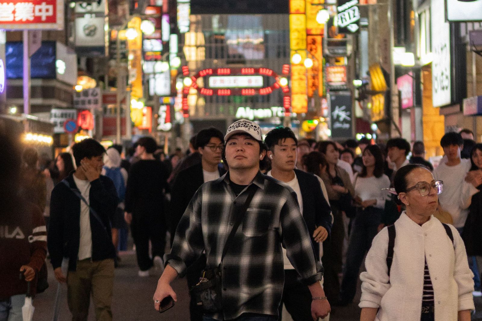 A crowded pedestrian street at night lit by dense neon signage, dozens of people walking past each other in different directions