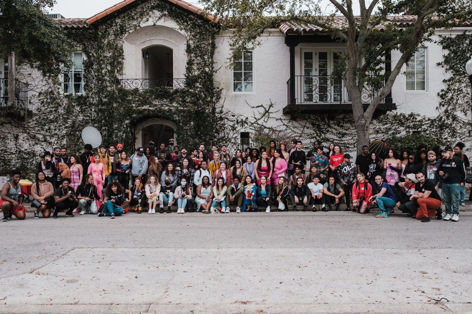 A very large group of around eighty people standing and sitting together in rows in front of an ivy-covered building for a group photo