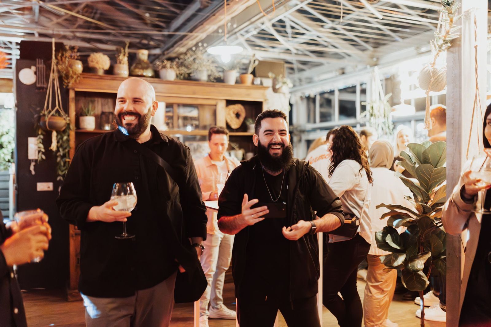 Two friends laughing together at an evening gathering indoors, one holding a phone, surrounded by other guests talking in small groups