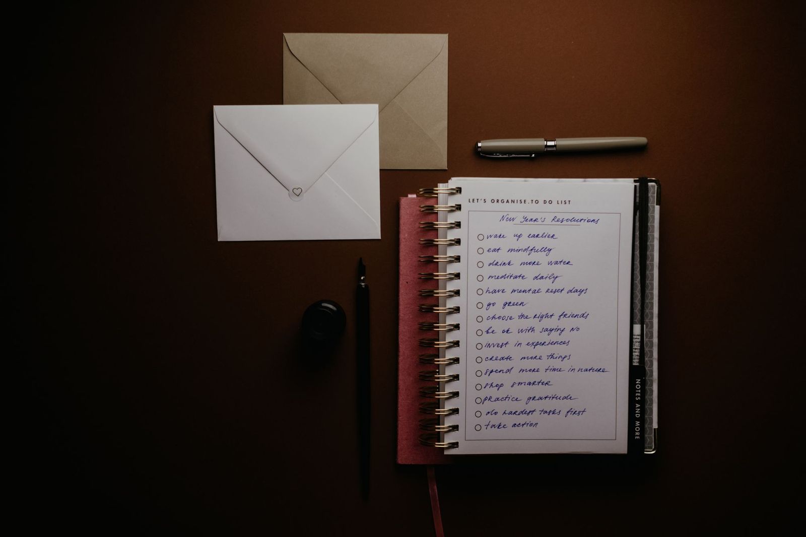 Two envelopes and a spiral notebook open to a handwritten checklist, laid out on a dark tabletop next to a pen and a bottle of ink