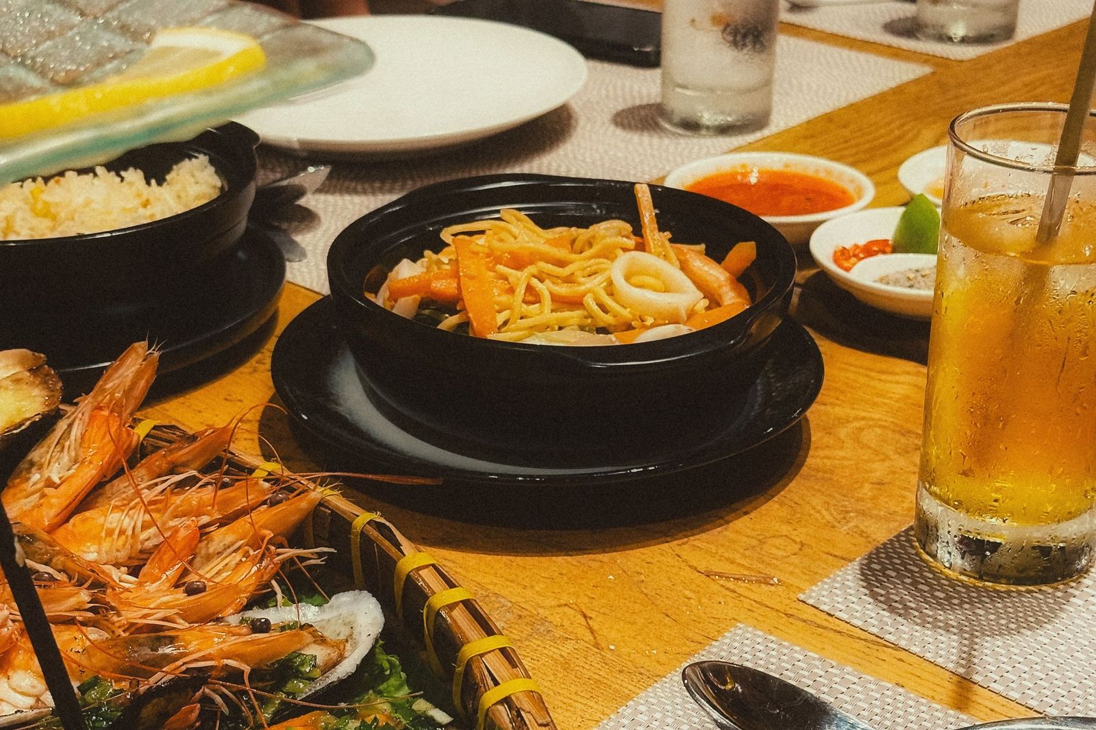 A table crowded with shared dishes at a Chinese seafood restaurant, including a hot claypot noodle dish, grilled prawns and oysters, and iced tea