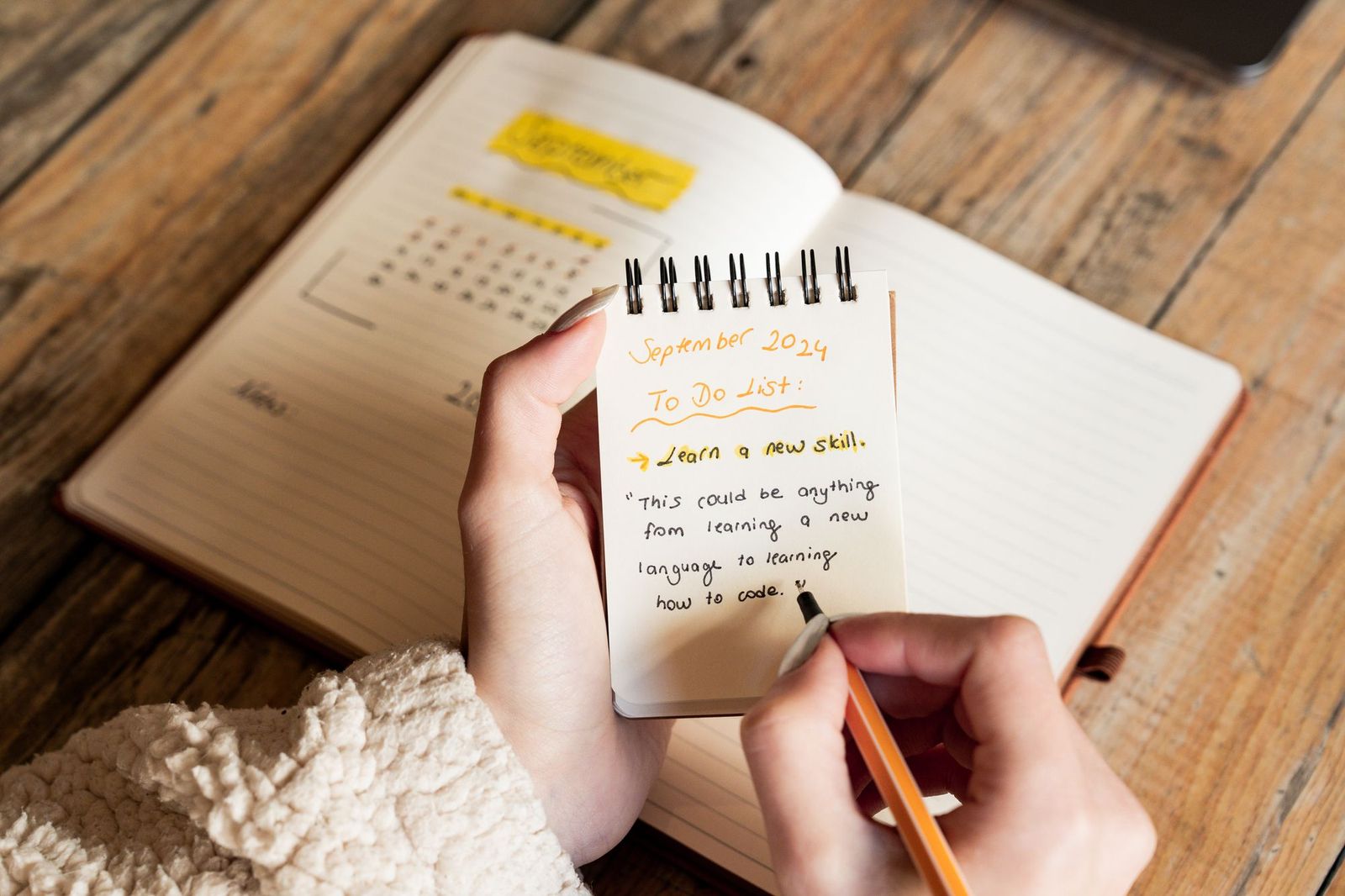 A person's hands writing a to-do list titled September 2024 in a small spiral notepad, resting on an open planner on a wooden desk