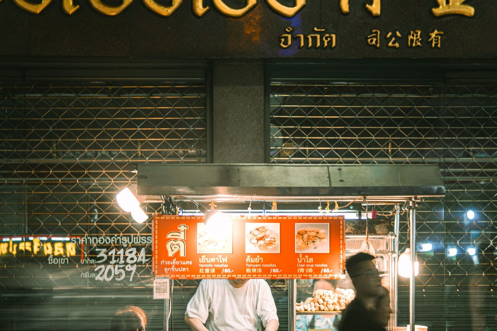 A brightly lit night market food stall in Thailand with an illuminated menu board showing dish photos and a QR code, a vendor working behind the counter