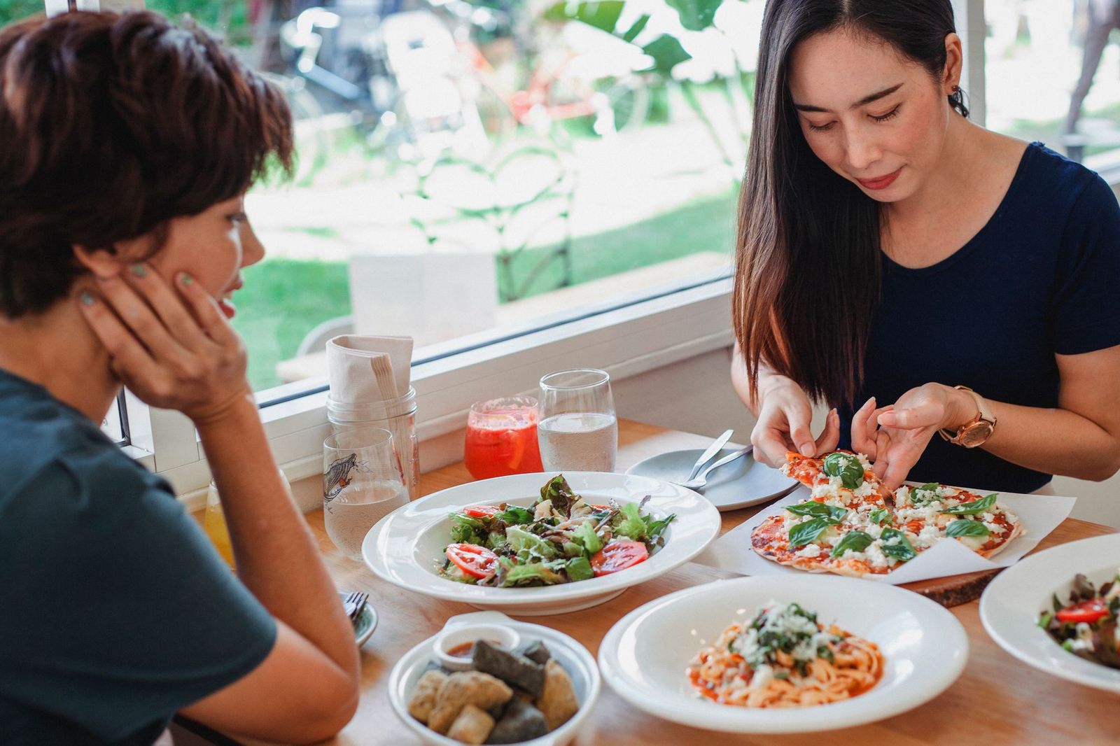 Two friends at a restaurant table leaning over shared plates of pizza, pasta and salad, mid conversation