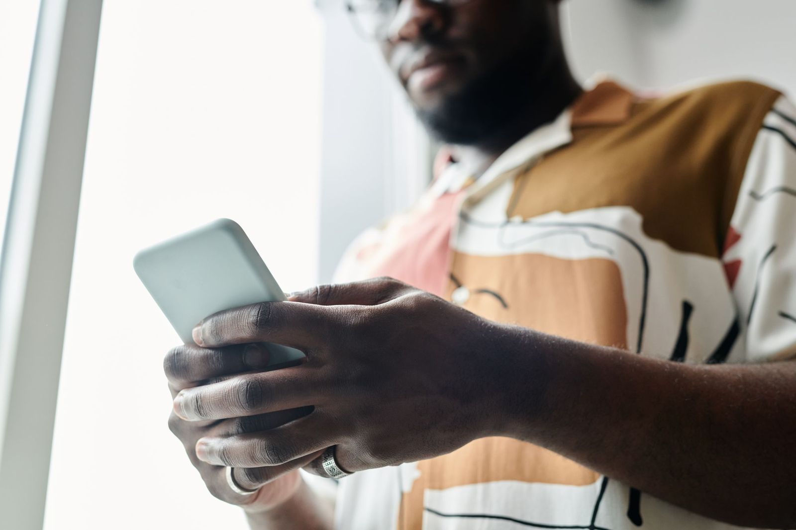 Close-up of a man's hands holding a phone in both hands by a bright window, thumb resting on the screen, his patterned shirt and glasses out of focus behind