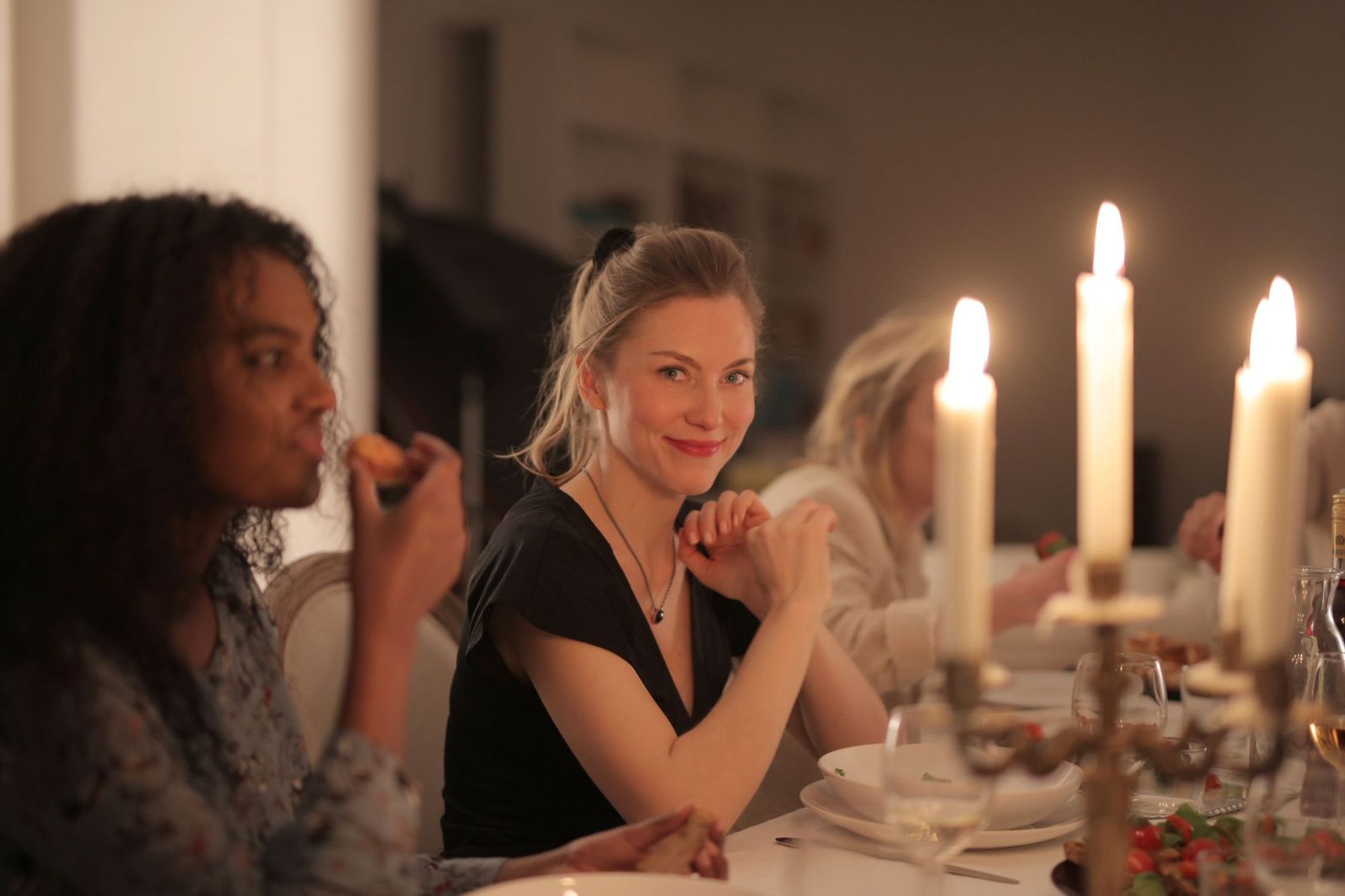 Several women sitting along one side of a long dinner table lit by tall candles, plates and glasses in front of them, one turning to smile at the camera