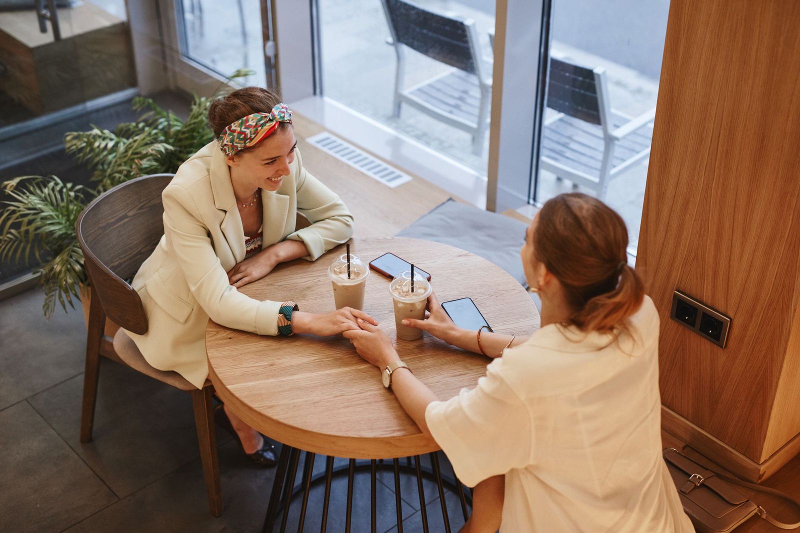 Two women sitting at a round wooden table in a bright cafe by a large window, iced coffees between them, phones on the table, holding hands and talking